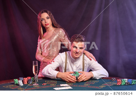 Man playing poker at casino, sitting at table with stacks of chips and cards on it. Celebrating win with woman. Black, smoke background. Close-up. Man playing poker at casino, sitting at table with stacks of chips and cards on it. Celebrating win with woman. Black, smoke background. Close-up. 87650614
