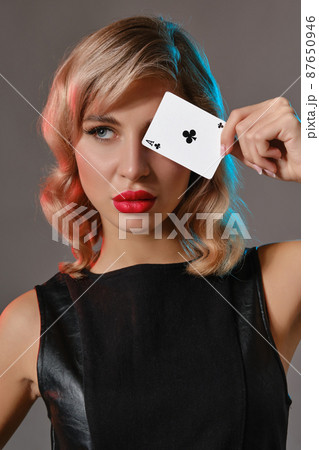Blonde girl in black leather dress showing ace of clubs, posing against gray background. Gambling entertainment, poker, casino. Close-up. Blonde girl in black leather dress showing ace of clubs, posing against gray background. Gambling entertainment, poker, casino. Close-up. 87650946