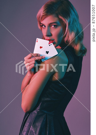 Blonde girl in black stylish dress showing two playing cards, posing against colorful background. Gambling entertainment, poker, casino. Close-up. Blonde girl in black stylish dress showing two playing cards, posing against colorful background. Gambling entertainment, poker, casino. Close-up. 87651007