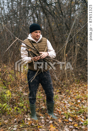 Vertical portrait of frozen tourist male wearing warm clothes collecting dry deadwood for fire in forest on overcast cold day. Concept of bushcraft, camping and survival in nature. 87651325