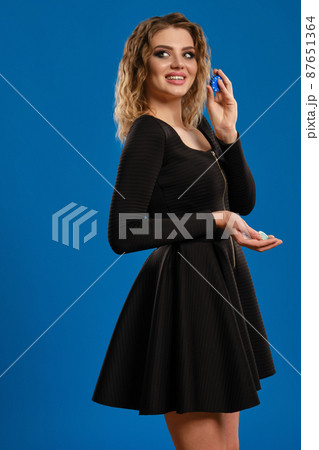 Blonde maiden in black short dress is smiling, showing colorful chips, posing sideways on blue background. Gambling, poker, casino. Close-up 87651364
