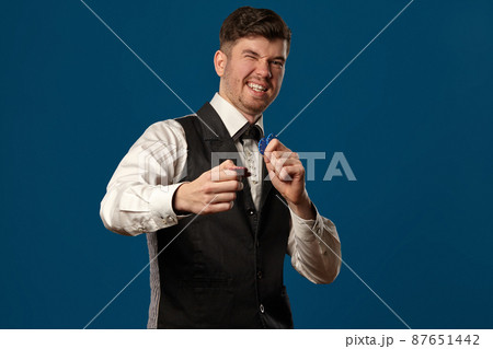 Newbie in poker, in black vest and white shirt. Holding some colored chips. Posing against blue background. Gambling, casino. Close-up. Newbie in poker, in black vest and white shirt. Holding some colored chips. Posing against blue background. Gambling, casino. Close-up. 87651442