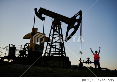 Petroleum operator in work vest raising hand while standing near pump jack. Oil man operating oil well while working in oil field in the evening. Concept of successful business. Petroleum operator in work vest raising hand while standing near pump jack. Oil man operating oil well while working in oil field in the evening. Concept of successful business. 87651595