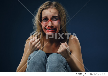 Frightened maiden in black t-shirt and jeans. Bleeding face covered with bruises, clenched her fists and crying, sitting on blue background. Close-up Frightened maiden in black t-shirt and jeans. Bleeding face covered with bruises, clenched her fists and crying, sitting on blue background. Close-up 87652233