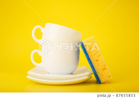 Dishwashing minimal concept. Plates, cups and cleaning sponge against bright yellow background Dishwashing minimal concept. Plates, cups and cleaning sponge against bright yellow background 87655853