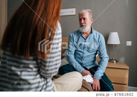 Back view of loving young granddaughter talking with gray-haired grandfather sitting in bed at cozy bedroom. Happy young woman and mature male having fun, enjoying leisure time, sharing news. Back view of loving young granddaughter talking with gray-haired grandfather sitting in bed at cozy bedroom. Happy young woman and mature male having fun, enjoying leisure time, sharing news. 87661602