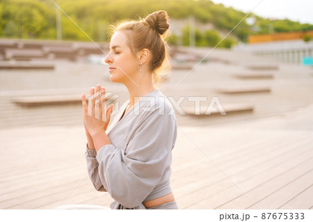 Side view of serene Caucasian young woman practicing yoga performing namaste pose with closed eyes outside in city park. Pretty calm female sitting lotus position on yoga mat outdoors alone. 87675333