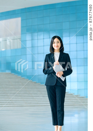 オフィスのエントランスにバソコンを持って立つ若いスーツ姿のビジネスウーマン 87677480