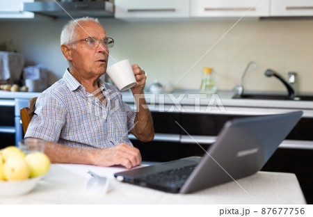 Old man sitting in kitchen at table with laptop 87677756