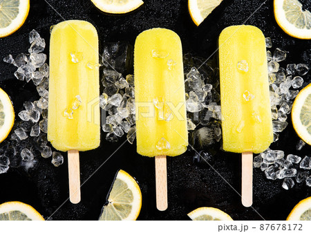 Three lemon popsicles on black background with ice and lemon slices. Flat lay composition. 87678172