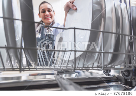 Happy young Caucasian woman removing or placing the dishes in the dishwasher in the kitchen of her apartment. Housework concept. Happy young Caucasian woman removing or placing the dishes in the dishwasher in the kitchen of her apartment. Housework concept. 87678186