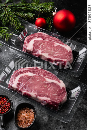 Rib-eye beef steak in plastic packing tray with Christmas tree decorations, on black dark stone table background 87692888