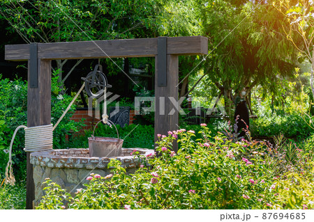 Beautiful artesian well made by stones and wheel pulley with metal bucket and rope in peaceful garden atmosphere. Retro stone water well in rural area. Garden decoration with antique items. 87694685