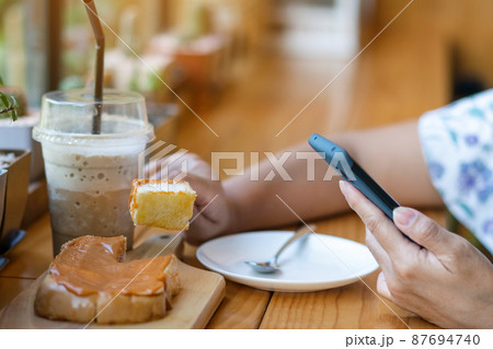 Close up to slices of toast bread and butter with steamed Thai tea custard topping in hand of woman using fork to eat for breakfast during using mobile phone. Relaxing with food and technology concept 87694740