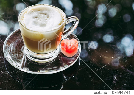 A red heart shaped candle on coasters with cappuccino coffee on scratched glass table. Candle in the form of a heart. Vintage, retro, old style. Image of Valentines day and love composition concept. A red heart shaped candle on coasters with cappuccino coffee on scratched glass table. Candle in the form of a heart. Vintage, retro, old style. Image of Valentines day and love composition concept. 87694741