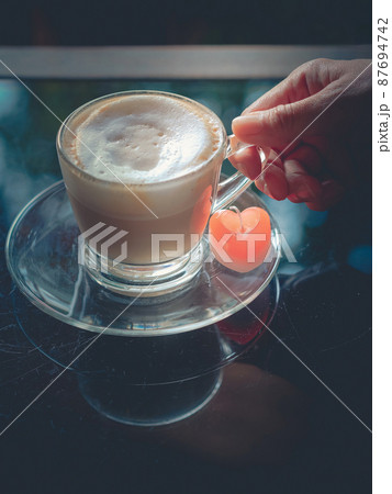 Hand of woman holding a cup of hot cappuccino coffee with red heart shaped candle on coasters on scratched glass table. Vintage,retro,old style. Image of Valentines day and love composition concept. 87694742