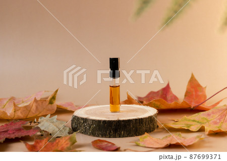 Glass perfume sample with brown liquid on a wooden tray standing on beige background with fallen leaves around. Luxury and natural autumn cosmetics presentation. Tester on a woodcut. Front view 87699371