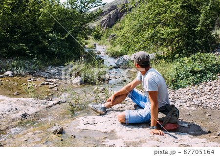 Young man traveler walking alone by a mountain river with rocky summer landscape Young man traveler walking alone by a mountain river with rocky summer landscape 87705164