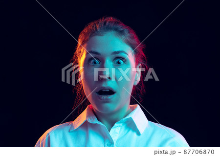 Shocked young girl in white shirt looking at camera with open mouth isolated on dark background in blue neon light, filter. Concept of emotions, fashion, youth Shocked young girl in white shirt looking at camera with open mouth isolated on dark background in blue neon light, filter. Concept of emotions, fashion, youth 87706870