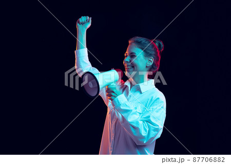 Half-length portrait of young pretty girl in white shirt shouting at megaphone isolated on dark background in blue neon light, filter. Concept of emotions, fashion, youth 87706882