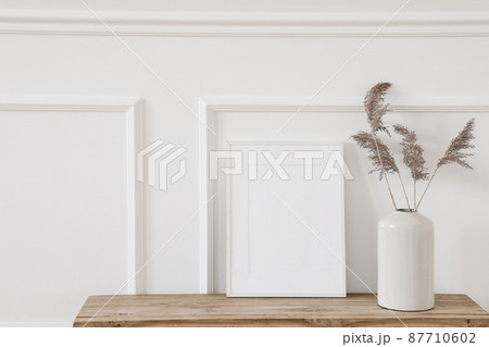 Blank white picture frame mockup. Vase with dry reed, grass on old wooden bench. Wall moulding background, trim decor. Elegant home interior decor, still life photo. Art dispaly. Front view. Blank white picture frame mockup. Vase with dry reed, grass on old wooden bench. Wall moulding background, trim decor. Elegant home interior decor, still life photo. Art dispaly. Front view. 87710602