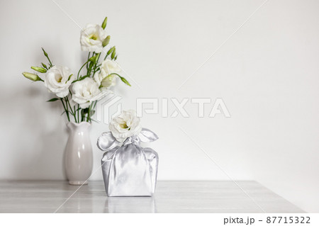 Gift box wrapped in silk fabric in Furoshiki technique, white flowers Eustoma or Lisianthus in vase on light wooden background. 87715322