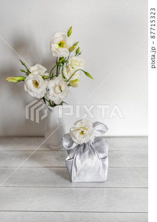 Gift box wrapped in silk fabric in Furoshiki technique, white flowers Eustoma or Lisianthus in vase on light wooden background. Gift box wrapped in silk fabric in Furoshiki technique, white flowers Eustoma or Lisianthus in vase on light wooden background. 87715323