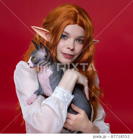 Portrait of young redhead woman cosplayer elf with little purebred Sphynx Hairless cat on red background. Elf in summer white dress hugging favorite kitten, smiling happiness and looking at camera. Portrait of young redhead woman cosplayer elf with little purebred Sphynx Hairless cat on red background. Elf in summer white dress hugging favorite kitten, smiling happiness and looking at camera. 87718552