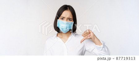 Portrait of asian woman in medical face mask showing thumbs down with disappointed, tired face expression, standing over white background Portrait of asian woman in medical face mask showing thumbs down with disappointed, tired face expression, standing over white background 87719313