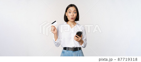 Online shopping concept. Image of surprised asian girl, holding credit card and smartphone, looking amazed in disbelief at camera, white background Online shopping concept. Image of surprised asian girl, holding credit card and smartphone, looking amazed in disbelief at camera, white background 87719318