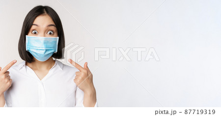 Image of amazed young asian woman pointing at herself while wearing medical face mask, standing over white background Image of amazed young asian woman pointing at herself while wearing medical face mask, standing over white background 87719319