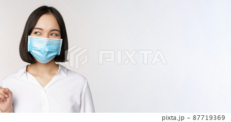 Portrait of cute asian woman in medical face mask, coronavirus protection, pointing finger left and looking intrigued at empty copy space, white background Portrait of cute asian woman in medical face mask, coronavirus protection, pointing finger left and looking intrigued at empty copy space, white background 87719369