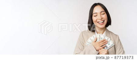 Happy asian businesswoman holding cash, hugging dollars money and smiling, standing over white background in suit Happy asian businesswoman holding cash, hugging dollars money and smiling, standing over white background in suit 87719373