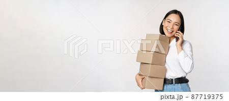 Image of young asian businesswoman answer phone call while carrying boxes for delivery, posing against white background Image of young asian businesswoman answer phone call while carrying boxes for delivery, posing against white background 87719375