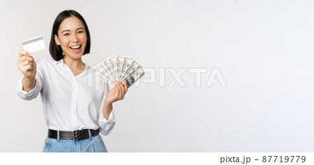 Happy korean woman holding credit card and money dollars, smiling and laughing, posing against white studio background Happy korean woman holding credit card and money dollars, smiling and laughing, posing against white studio background 87719779