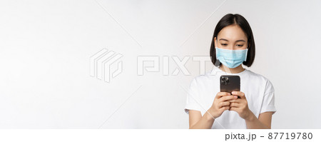 Health, covid and mobile concept. Young asian woman in medical face mask, looking at smartphone screen, using phone app, shopping online, white background Health, covid and mobile concept. Young asian woman in medical face mask, looking at smartphone screen, using phone app, shopping online, white background 87719780