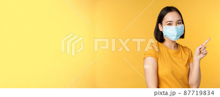 Vaccination from covid and health concept. Image of smiling korean girl in medical face mask, band aid on shoulder, pointing finger at banner advertisement, yellow background 87719834