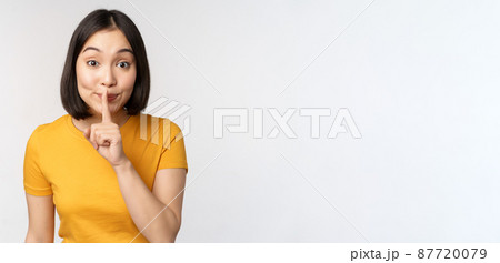 Keep quiet. Cute asian woman make shhh gest, showing shush, hush sign, press finger to lips, silence, standing over white background 87720079