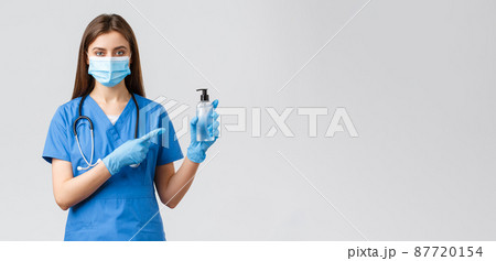 Covid-19, preventing virus, health, healthcare workers and quarantine concept. Young professional female nurse or doctor in blue scrubs, medical mask, pointing finger at hand sanitizer Covid-19, preventing virus, health, healthcare workers and quarantine concept. Young professional female nurse or doctor in blue scrubs, medical mask, pointing finger at hand sanitizer 87720154