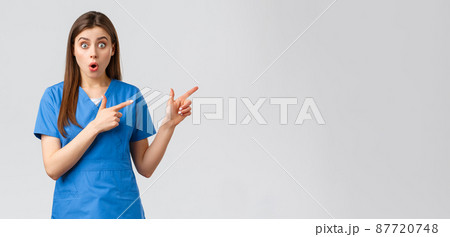 Healthcare workers, prevent virus, insurance and medicine concept. Surprised nurse or doctor in blue scrubs listen to interesting news, pointing fingers right, gasping shocked at camera 87720748