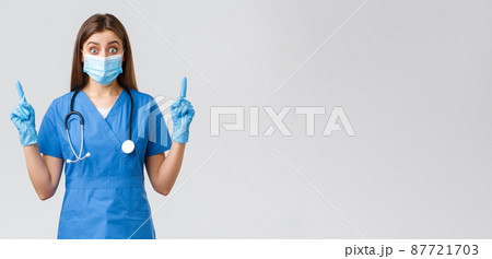 Covid-19, preventing virus, health, healthcare workers concept. Surprised or curious female nurse in blue scrubs, personal protective equipment, pointing fingers up, found out interesting news Covid-19, preventing virus, health, healthcare workers concept. Surprised or curious female nurse in blue scrubs, personal protective equipment, pointing fingers up, found out interesting news 87721703