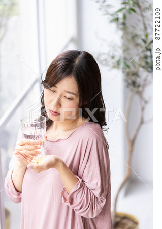 カプセル錠剤とグラスを持って不安げな表情を見せる部屋着の女性 カプセル錠剤とグラスを持って不安げな表情を見せる部屋着の女性 87722109