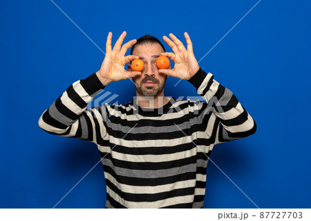 Portrait of a funny man dressed in a striped sweater holding some small oranges in front of his eyes simulating glasses. Fun concept 87727703