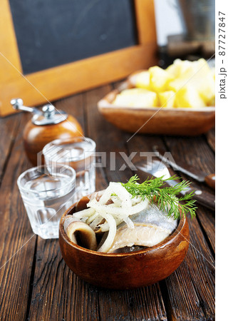 bowl of salted herring on the background of a glass of vodka and decanter. Salted herring in jar as a snack for vodka. bowl of salted herring on the background of a glass of vodka and decanter. Salted herring in jar as a snack for vodka. 87727847