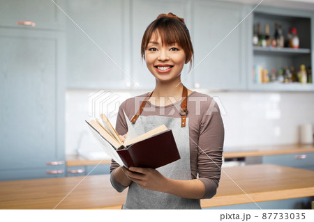 smiling woman in apron with cook book at kitchen 87733035