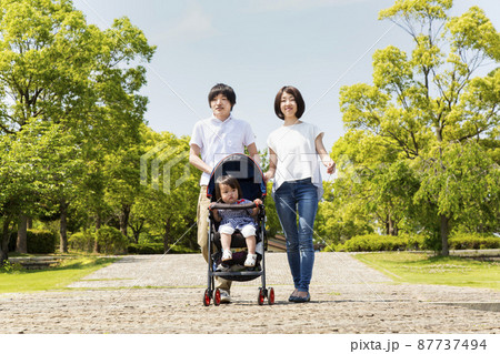 新緑の公園の遊歩道をベビーカーを押しながら散歩する幸せな夫婦。家族,幸せ,愛情のイメージ 87737494