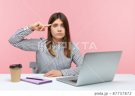 Upset woman office worker showing stupid gesture with finger looking at camera with displeased tired expression, sitting at workplace with laptop. Indoor studio shot isolated on pink background 87737872