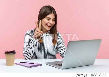 Successful positive woman office worker showing victory gesture and smiling looking into laptop screen, talking video call, online conference. Indoor studio shot isolated on pink background Successful positive woman office worker showing victory gesture and smiling looking into laptop screen, talking video call, online conference. Indoor studio shot isolated on pink background 87737876