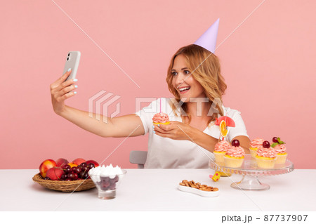 Smiling blonde woman celebrating birthday online with followers, broadcasting livestream, showing tasty cake with candle to camera, looks happy. Indoor studio shot isolated on pink background. Smiling blonde woman celebrating birthday online with followers, broadcasting livestream, showing tasty cake with candle to camera, looks happy. Indoor studio shot isolated on pink background. 87737907