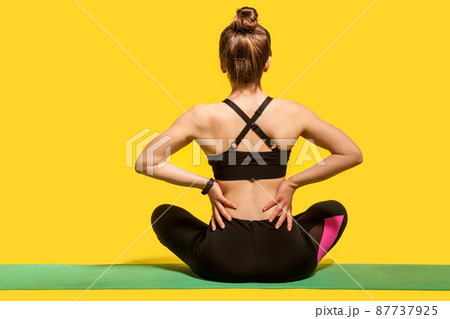 Back view, sportswoman sitting on mat and touching sore spine, suffering backache after physical training or yoga practice, massaging stiff muscles. indoor studio shot isolated on yellow background Back view, sportswoman sitting on mat and touching sore spine, suffering backache after physical training or yoga practice, massaging stiff muscles. indoor studio shot isolated on yellow background 87737925